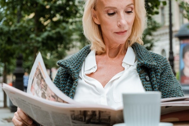 A tenant with a blond bob haircut sits at an outdoor cafe and reviews for rent ads in the classified section of a newspaper