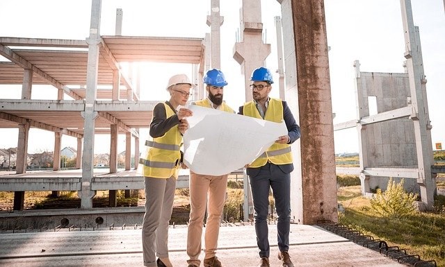 Three construction workers in yellow vests and hardhats stand in front of a partially built development and review a building plan